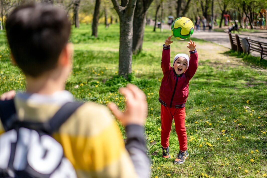 LBV-032 Ein kleines Kind in roter Kleidung wirft mit erhobenen Armen lachend einen Ball in Richtung einer Person im Vordergrund, die nur von hinten zu sehen ist. Die Szene spielt in einem sonnigen Park.