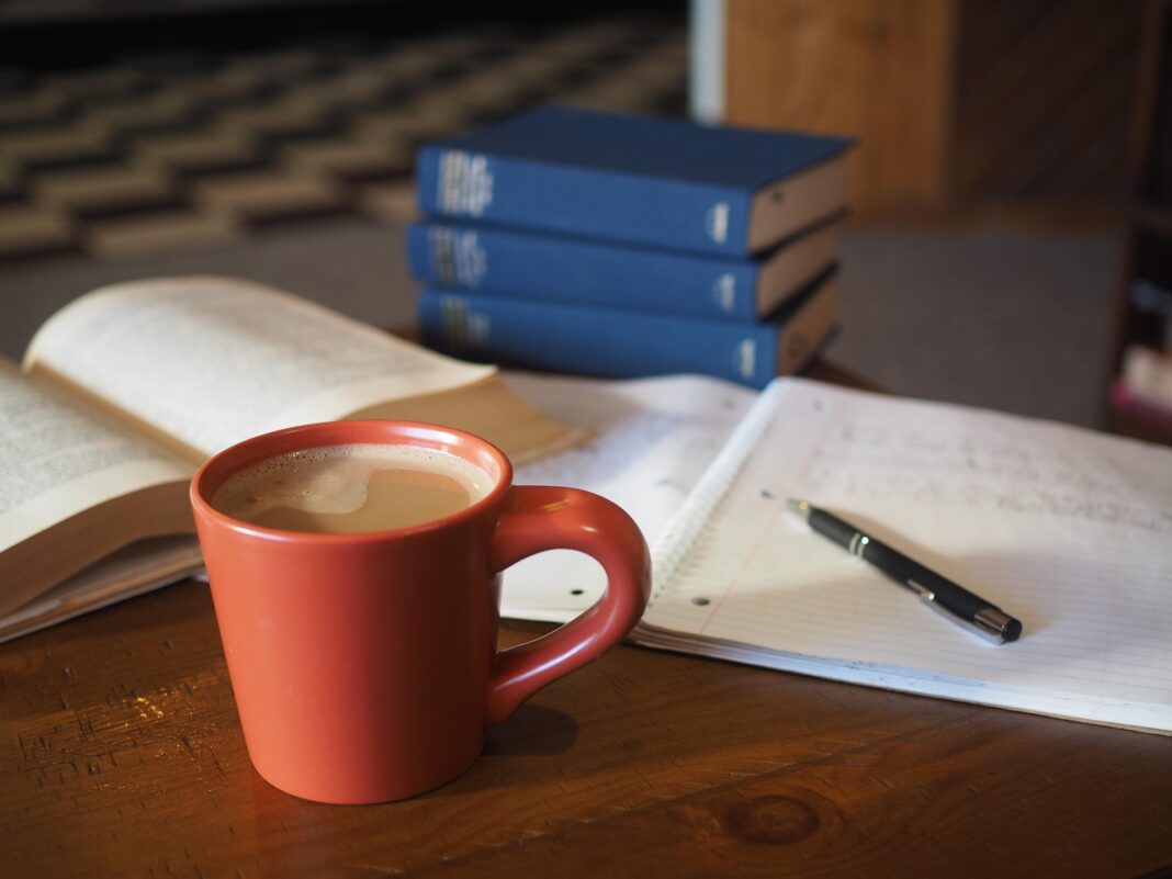 LBV-041 Orangefarbene Kaffeetasse auf einem Holztisch neben einem offenen Buch, Notizzettel und Kugelschreiber, mit mehreren blauen Ordnern im Hintergrund – eine ruhige Lernsituation.