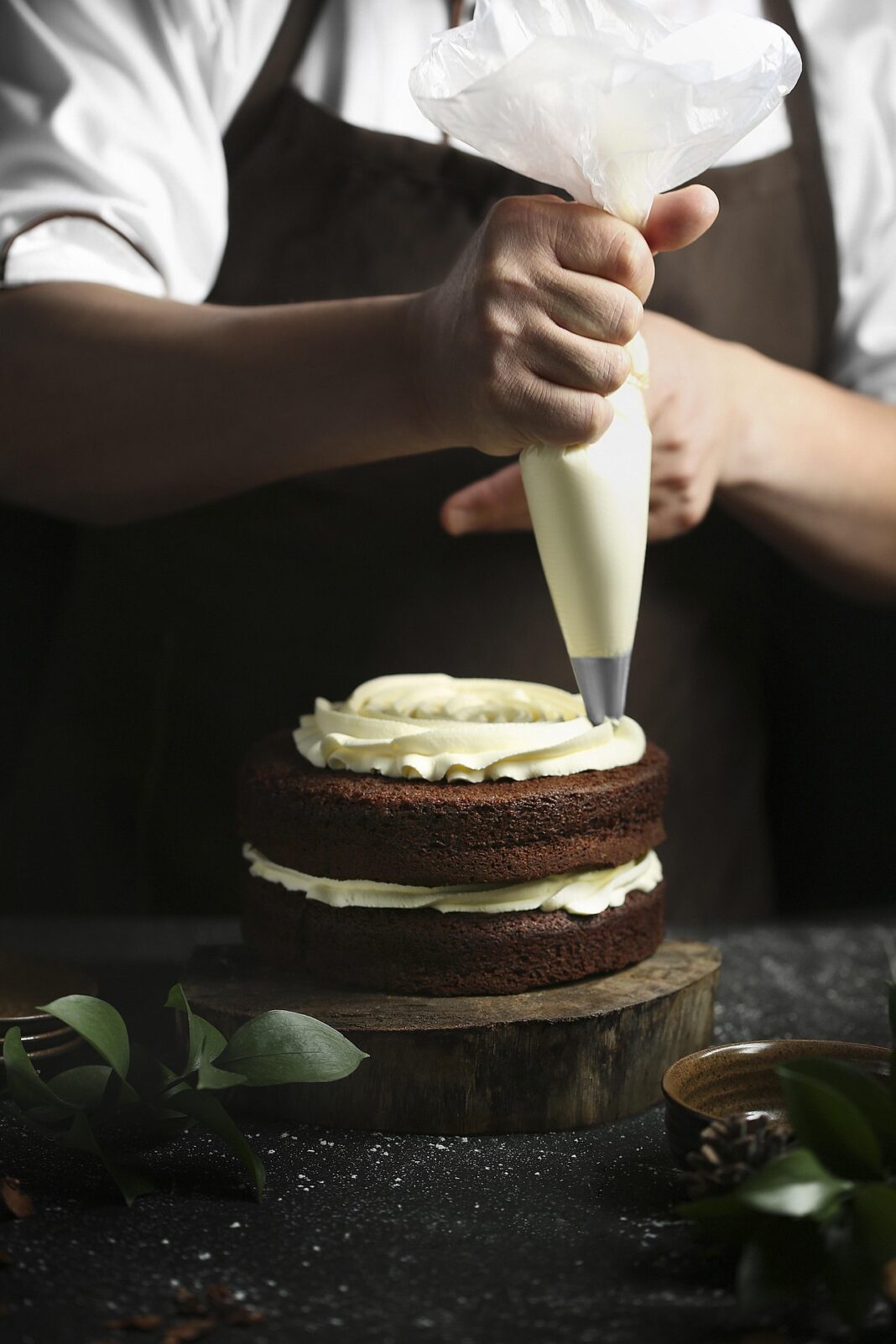 Eine Person mit weißem Hemd und brauner Schürze dekoriert eine zweischichtige Schokoladentorte mit einer Spritztülle und heller Buttercreme.