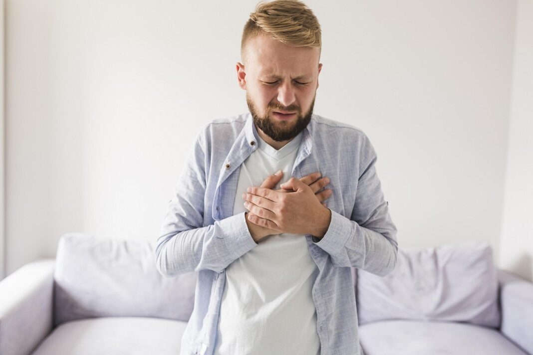 heartburn-5273873_1920 Ein junger Mann mit blondem Bart hält sich mit schmerzverzerrtem Gesicht die Brust – er steht im Wohnzimmer und scheint unter Sodbrennen zu leiden.