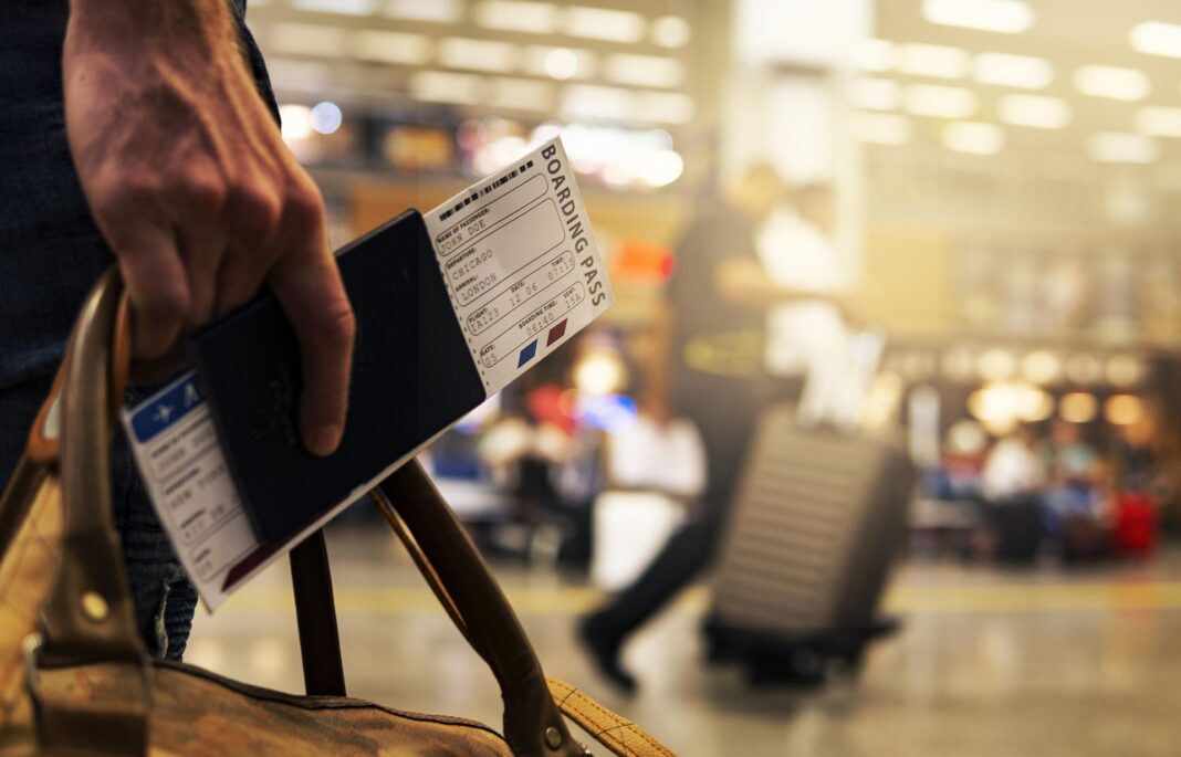 Nahaufnahme einer Hand, die eine Reisetasche trägt, mit einem Reisepass und einer Bordkarte für einen Flug von Chicago nach London; im Hintergrund verschwommen Reisende mit Rollkoffern in einem Flughafen.