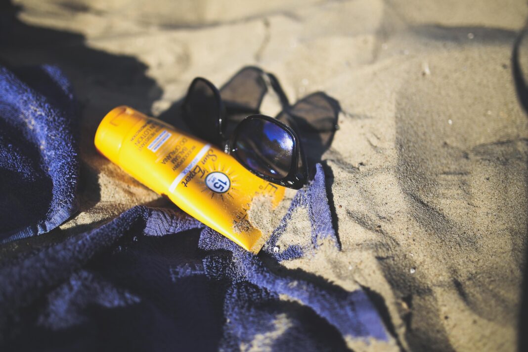 sun-791524_1920 Sonnencreme, Sonnenbrille und blaues Handtuch liegen auf sandigem Strandboden.