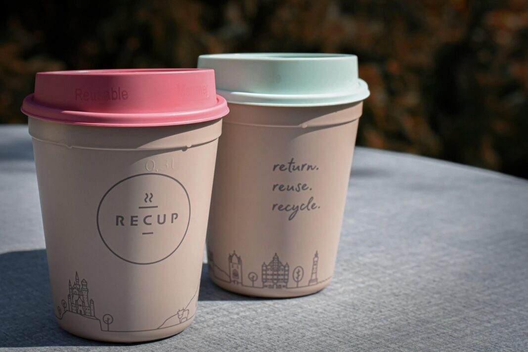 Zwei Mehrwegbecher aus Kunststoff mit unterschiedlich farbigen Deckeln – einer rosa, der andere mintgrün – stehen auf einem Tisch im Freien. Auf dem vorderen Becher steht das Logo „RECUP“, auf dem hinteren der Schriftzug „return. reuse. recycle.“