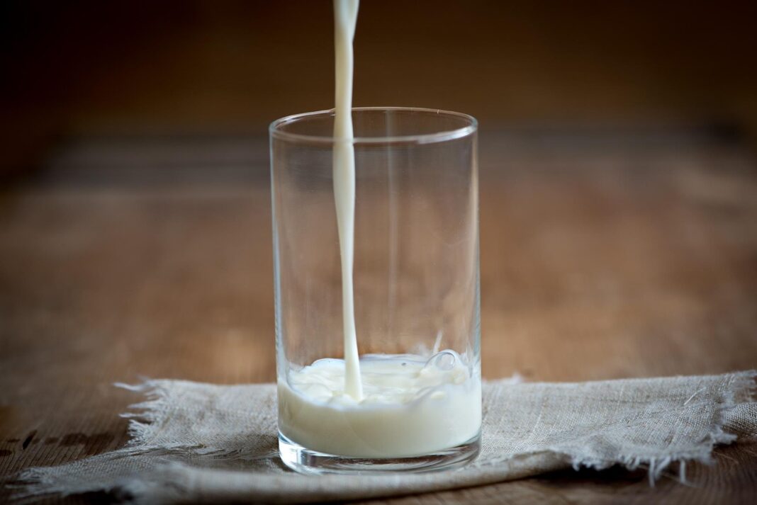 Ein klarer Glasbecher, in den gerade Milch gegossen wird, steht auf einem groben Stofftuch auf einem Holztisch.