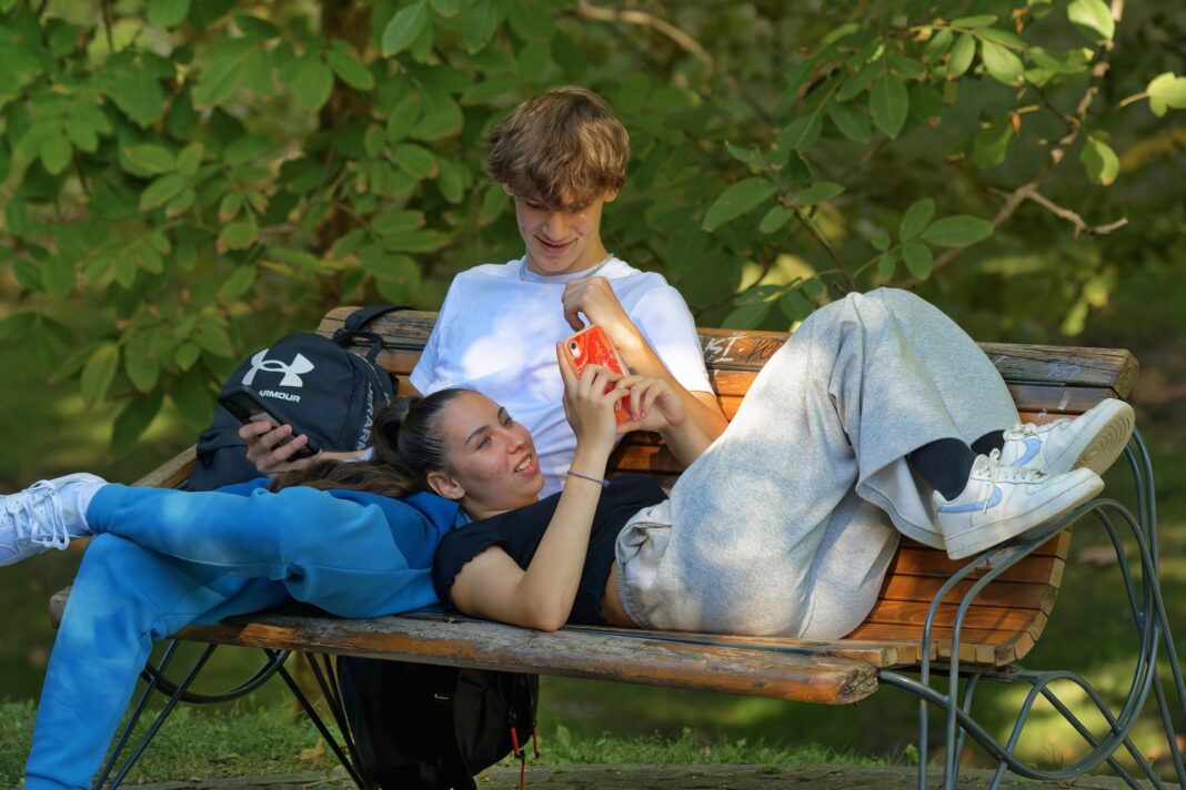 couple-7481134_1920 Zwei Jugendliche liegen entspannt auf einer Parkbank. Ein Junge in weißem T-Shirt sitzt aufrecht, während ein Mädchen in grauer Jogginghose und schwarzem Shirt mit dem Kopf auf seinem Schoß liegt. Beide schauen gemeinsam auf ein Smartphone. Neben ihnen liegt ein schwarzer Rucksack. Im Hintergrund ist grünes Laub sichtbar.