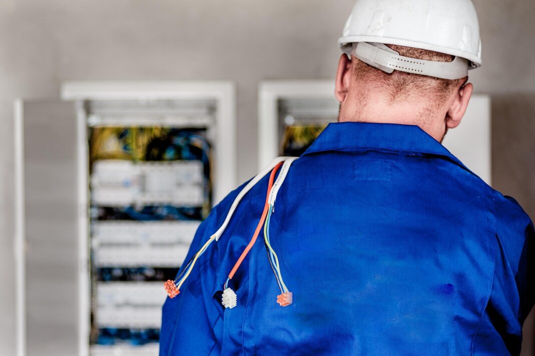 electrician-1080586_1920 Elektriker in blauer Arbeitskleidung und Schutzhelm steht mit dem Rücken zur Kamera, an dessen Schulter mehrere elektrische Kabel hängen; im Hintergrund sind geöffnete Stromverteiler zu sehen.