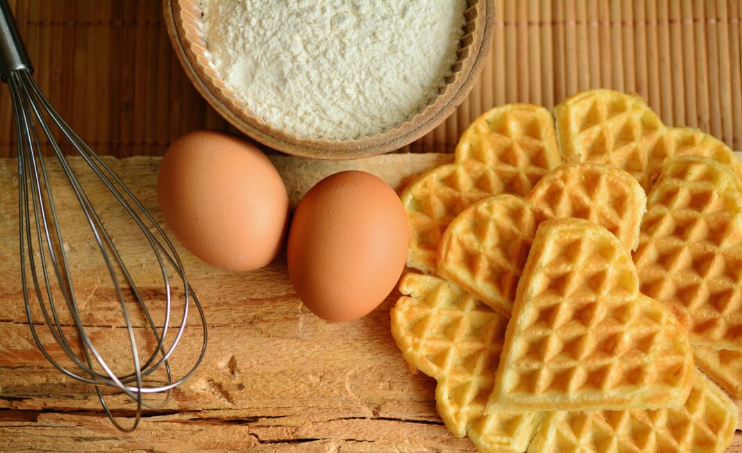 Zutaten für Waffeln: Zwei braune Eier, eine Schüssel mit Mehl, ein Schneebesen und mehrere goldbraune Herzwaffeln auf einer Holzunterlage.