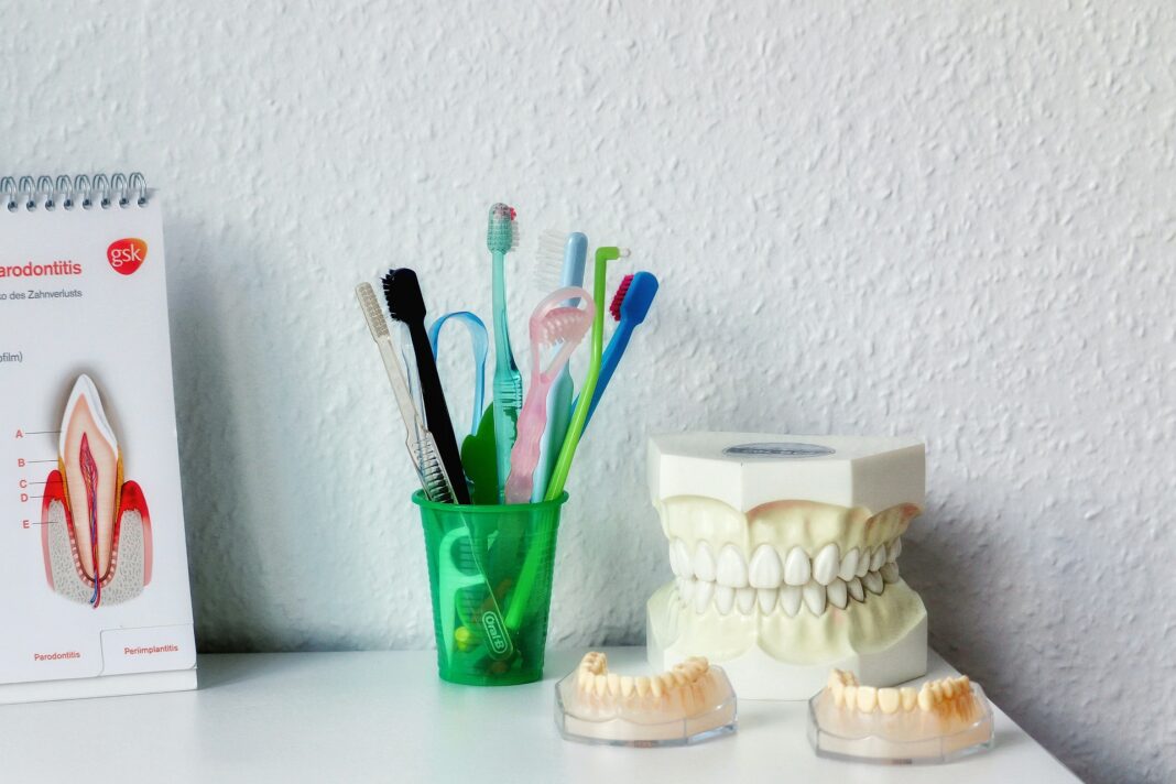 Verschiedene Zahnbürsten in einem grünen Plastikbecher neben einem Zahnmodell und Informationsmaterial über Parodontitis auf einem weißen Tisch.