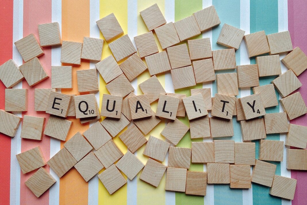 LBV-089 Holz-Spielsteine auf einem bunten Regenbogen-Hintergrund formen das Wort „EQUALITY“ (Gleichheit).