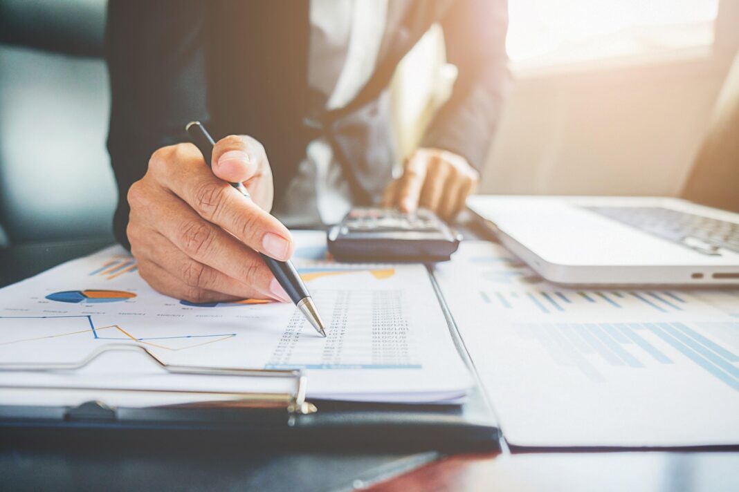 finance-7529921_1920 Person in Business-Outfit analysiert Finanzdokumente mit Diagrammen und Zahlen, nutzt Taschenrechner und Laptop am Schreibtisch.