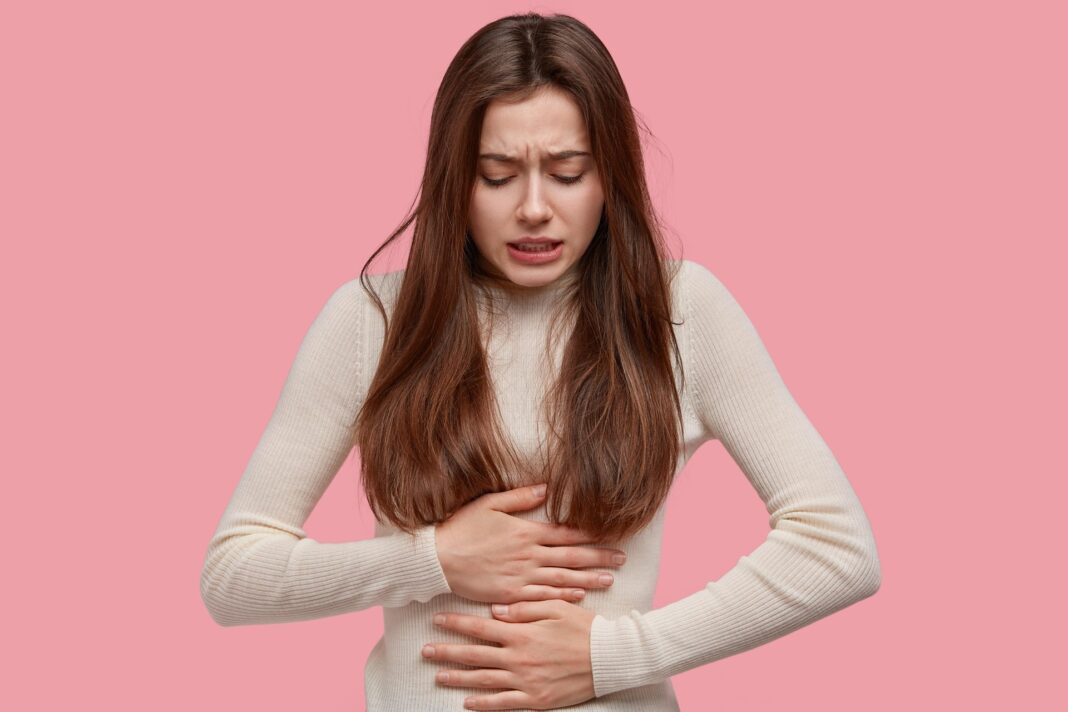 Junge Frau mit langem braunem Haar fasst sich mit schmerzverzerrtem Gesicht an den Bauch, vor rosafarbenem Hintergrund.