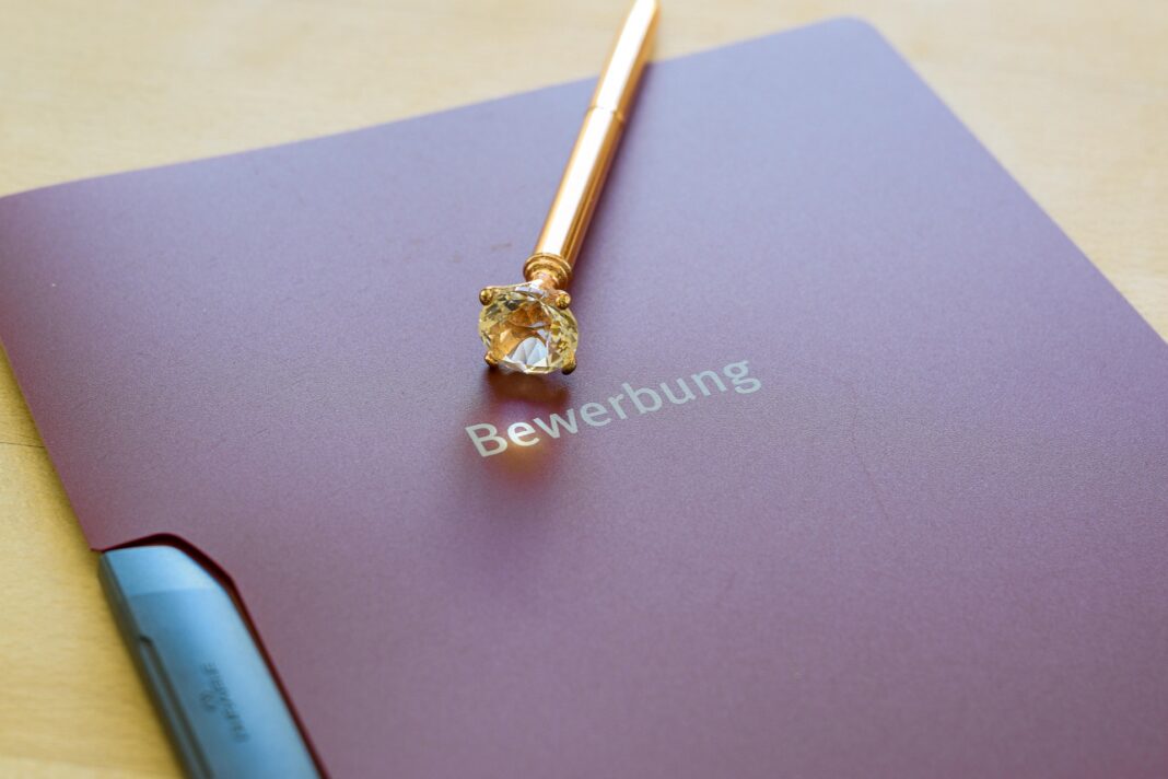 Eine weinrote Bewerbungsmappe mit der Aufschrift „Bewerbung“ liegt auf einem Holztisch. Darauf befindet sich ein goldfarbener Kugelschreiber mit einem großen, funkelnden Schmuckstein an der Spitze.